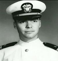 A black and white head shot of Brian from his Navy days. He's in his dress whites and looking to the right of camera.