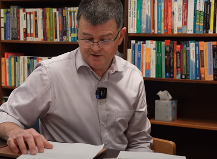 Professor Bradley Dilger sits at a desk in front of a bookshelf. He is speaking and pointing to a piece of paper on his desk.