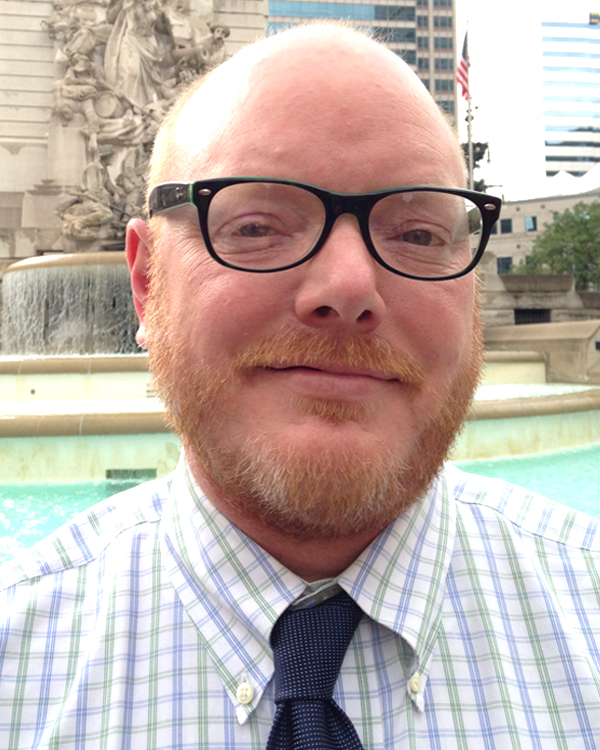 Headshot of Dr. Denny smiling at the camera with a fountain behind him.