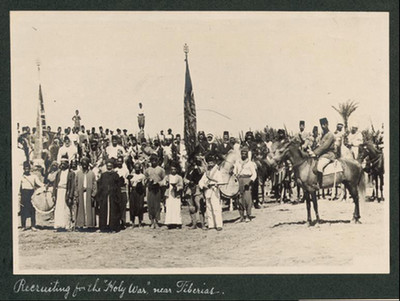 A black-and-white historical photograph titled "Recruiting for the Holy War near Tiberias." It depicts a large group of Ottoman soldiers and recruits gathered in a desert clearing, some on horseback and others standing, with several tall flags held upright throughout the crowd.