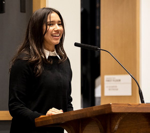 Johari Tello speaking at a department lecture with event presentation slides visible in the background"
