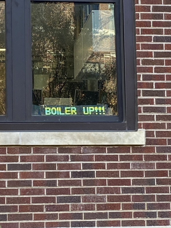 An LCD sign with the phrase "Boiler Up" sitting in the window of a red brick building.