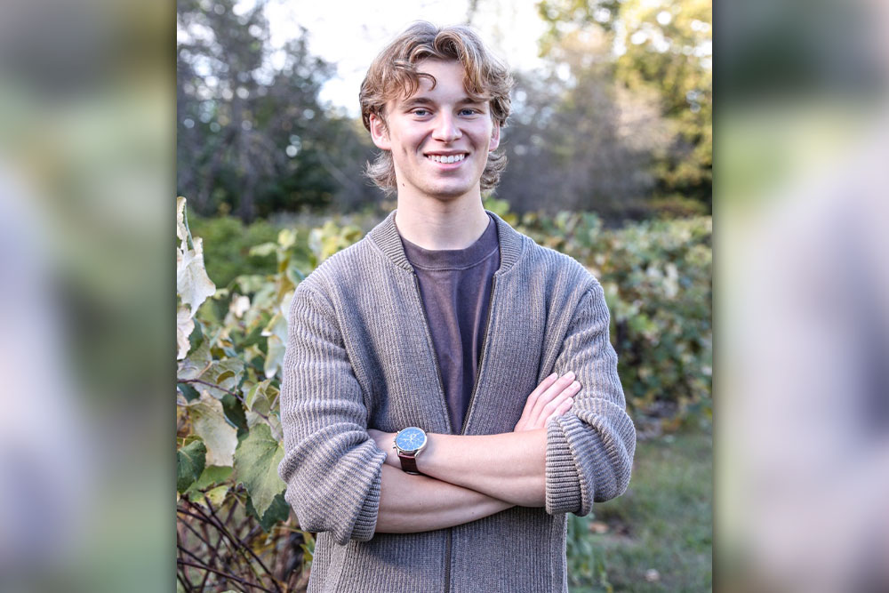 “Person standing outdoors with arms crossed, wearing a cardigan in front of leafy plants and trees.”