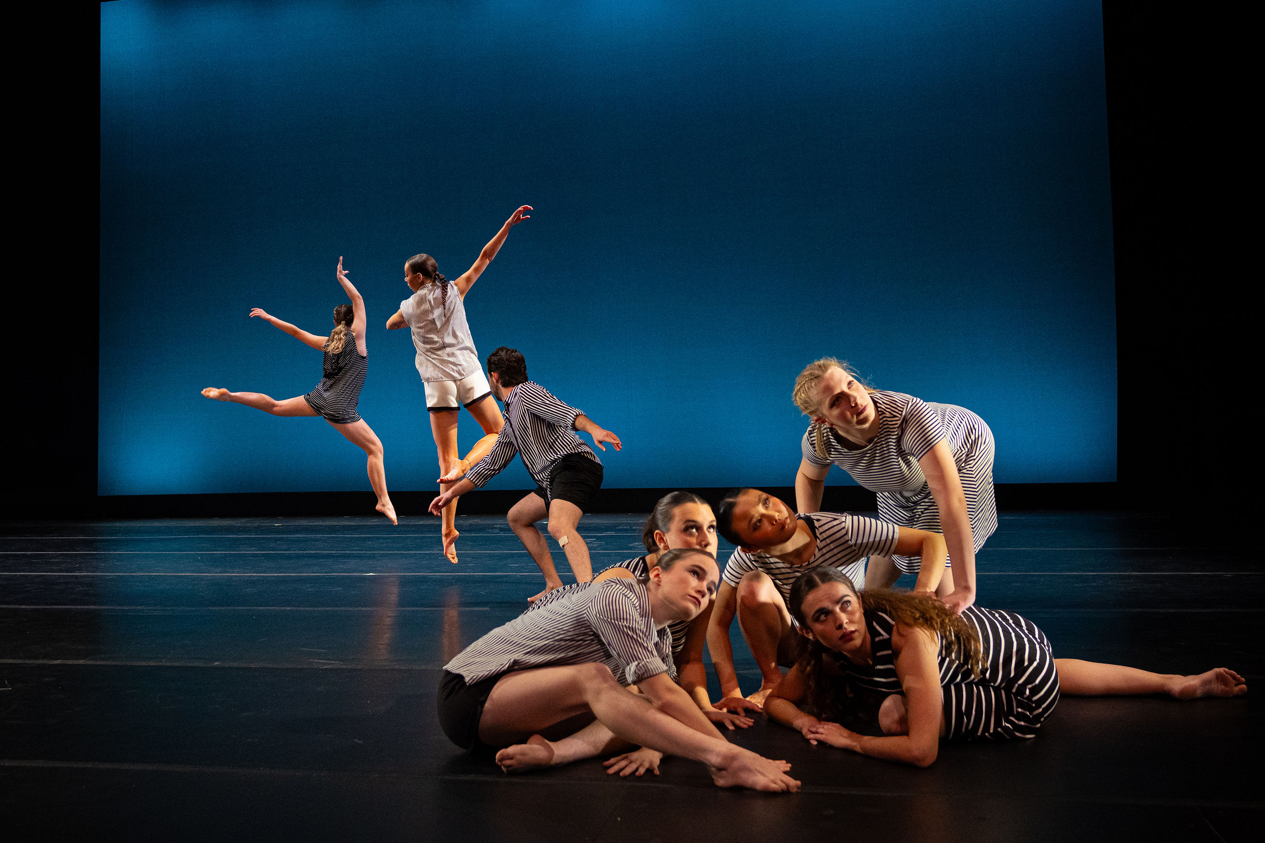 A cluster of dancers close to the camera looking with curiosity in front of a blue background. Behind them, three dancers leaping.