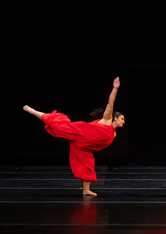 A dancer in red on a black background with one arm pointed up and one arm pointed behind.