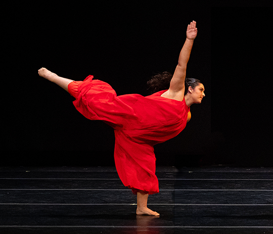 A dancer in red on a black background with one arm pointed up and one arm pointed behind.