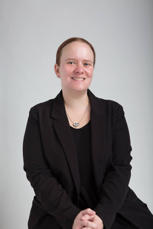 A headshot of Elise Taylor. She's wearing all black and smiling at the camera against a neutral background. 