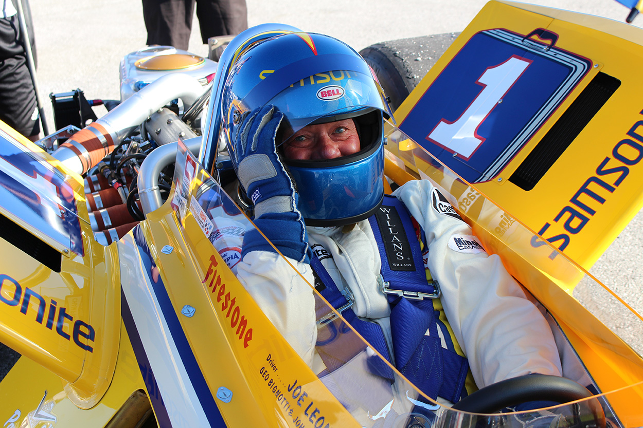 Chuck Jones wearing a full race suit and helmet sitting inside of a yellow Indy Car.