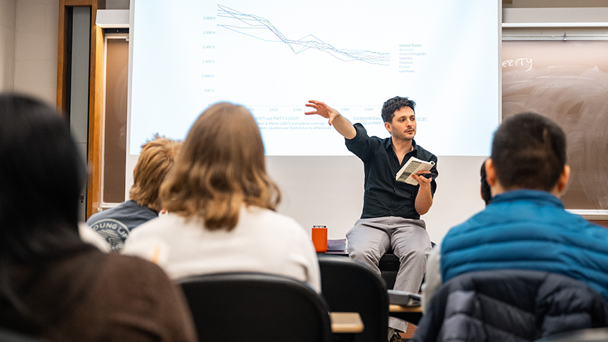 JP Messina, sitting on a table facing students, reads from a book. A graph is projected on the screen behind him.