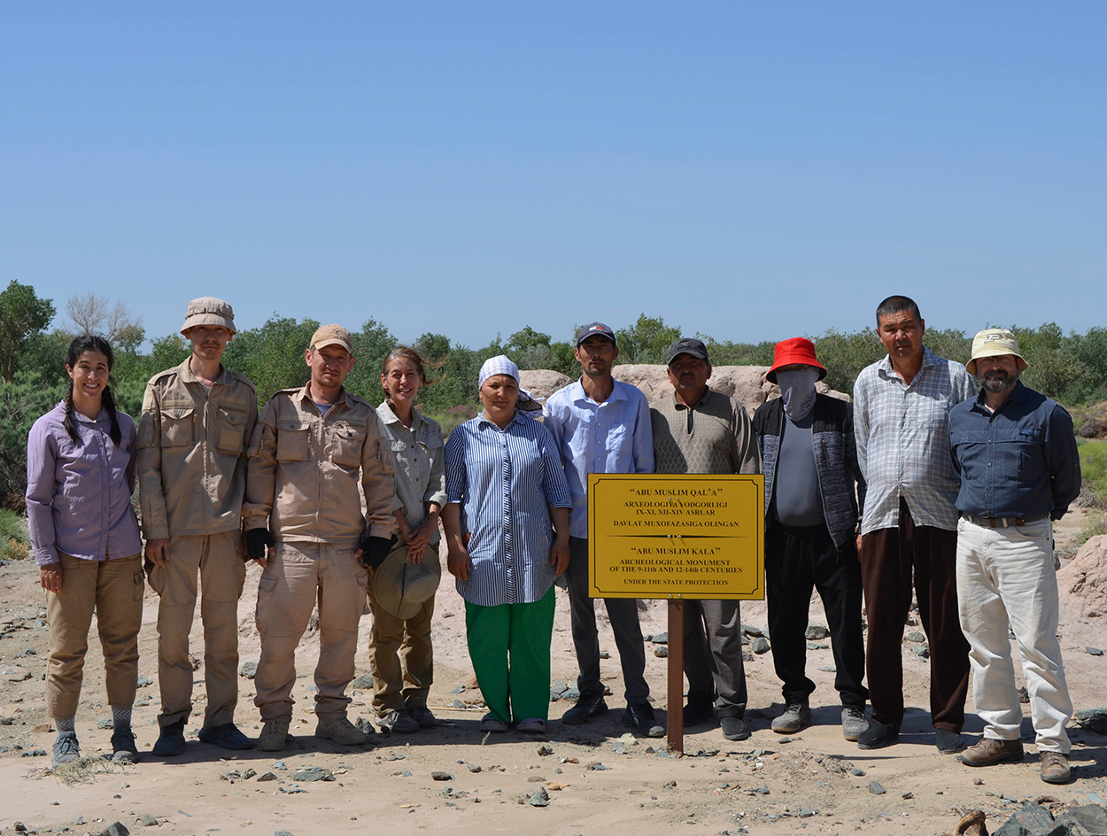 A group of ten people standing with a sign reading “Abu Muslim Qal’a”