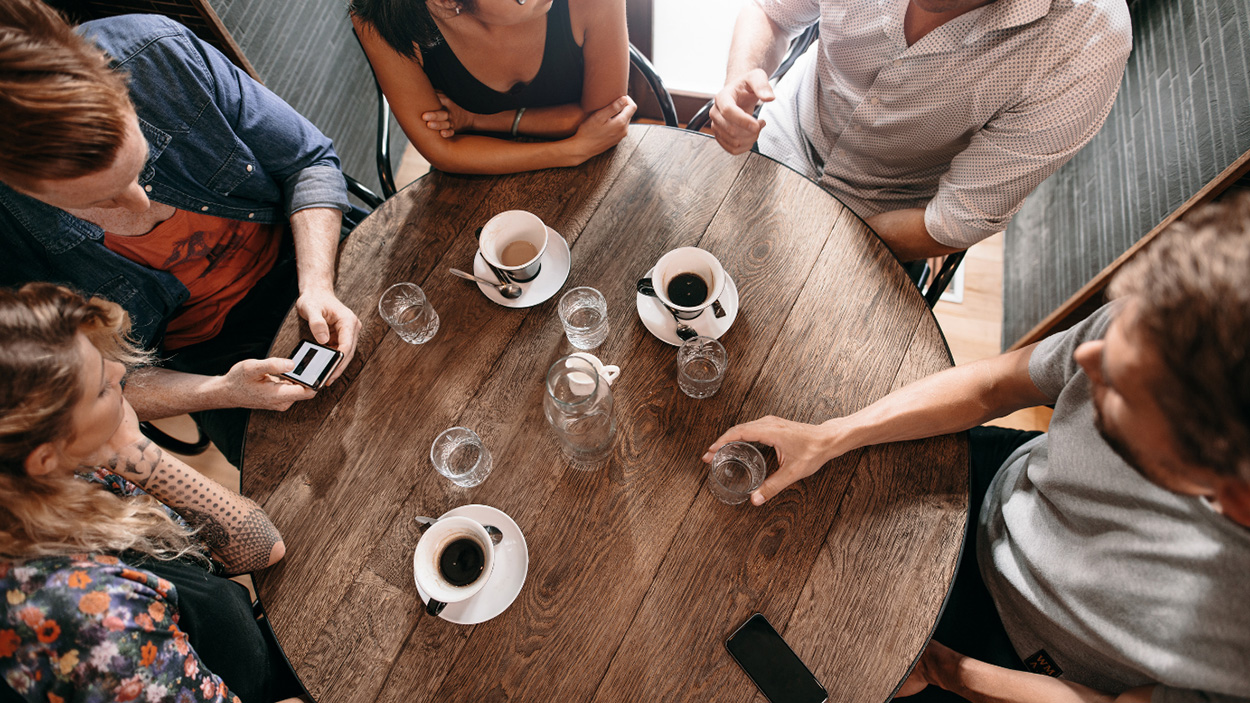 Five people sitting around a round wood table drinking water and coffee.