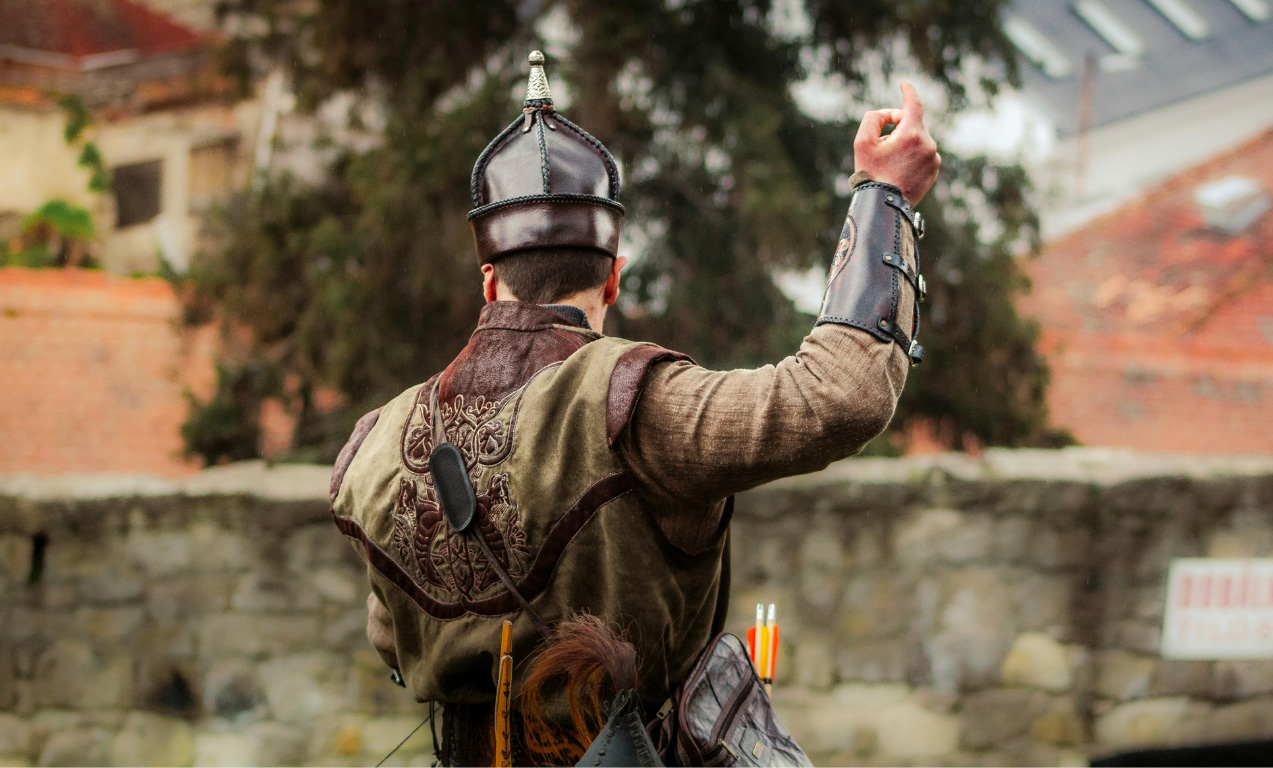 A person viewed from behind, dressed in historical medieval-style attire, including a dark leather helmet with a silver-topped peak, a brown patterned vest, and a leather bracer on their raised left arm. They are standing in front of a low stone wall, with arrows visible in a quiver at their hip.