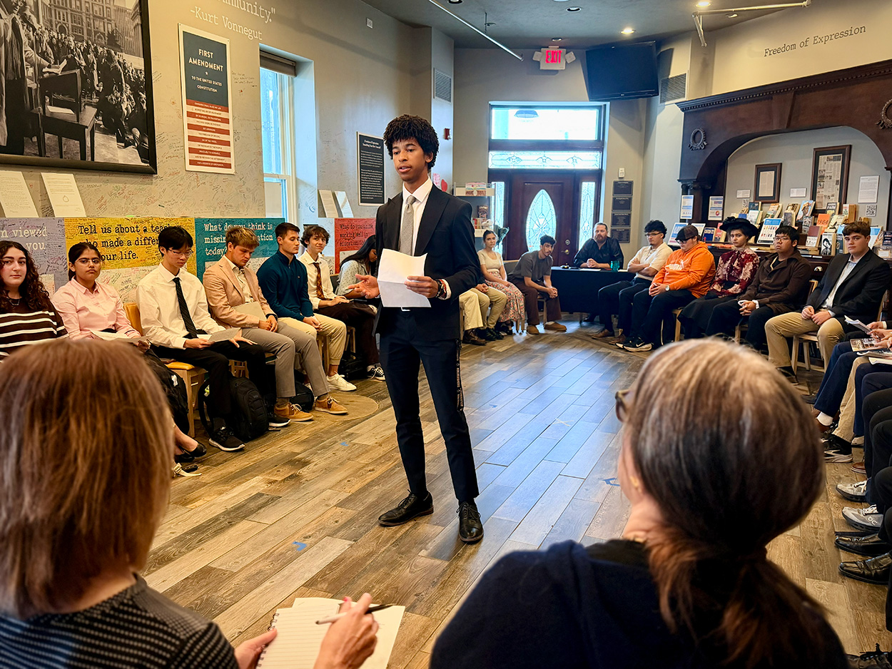 A student in a suit standing in a crowded room for Frankenstein’s trial.