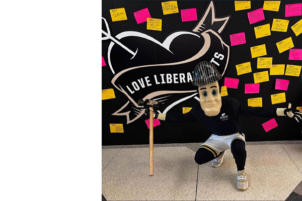 Purdue Pete kneeling in front of a College of Liberal Arts sign