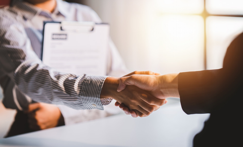 Two people shaking hands with a clipboard holding a resume in the background